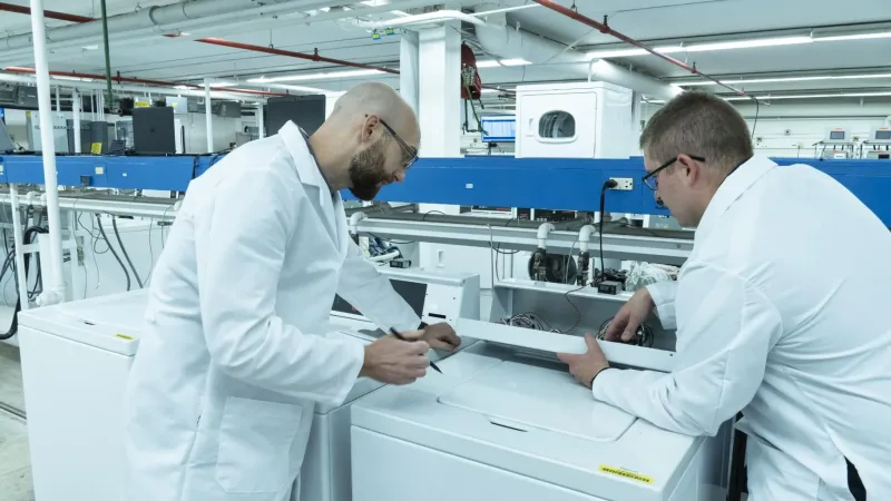Two men in white lab coats working on washing machine in test lab
